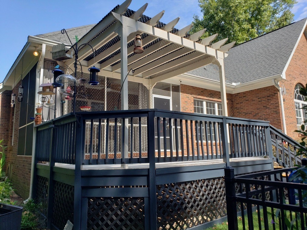 Wooden pergola built over a raised backyard deck with dark railing and hanging lights behind a brick home.