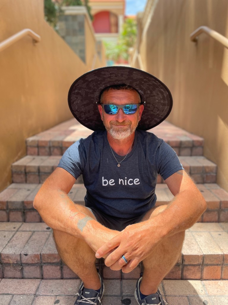 David, owner of David's Home Solutions, is sitting on a step outside with glasses and a hat on and a t-shirt that says "be nice".